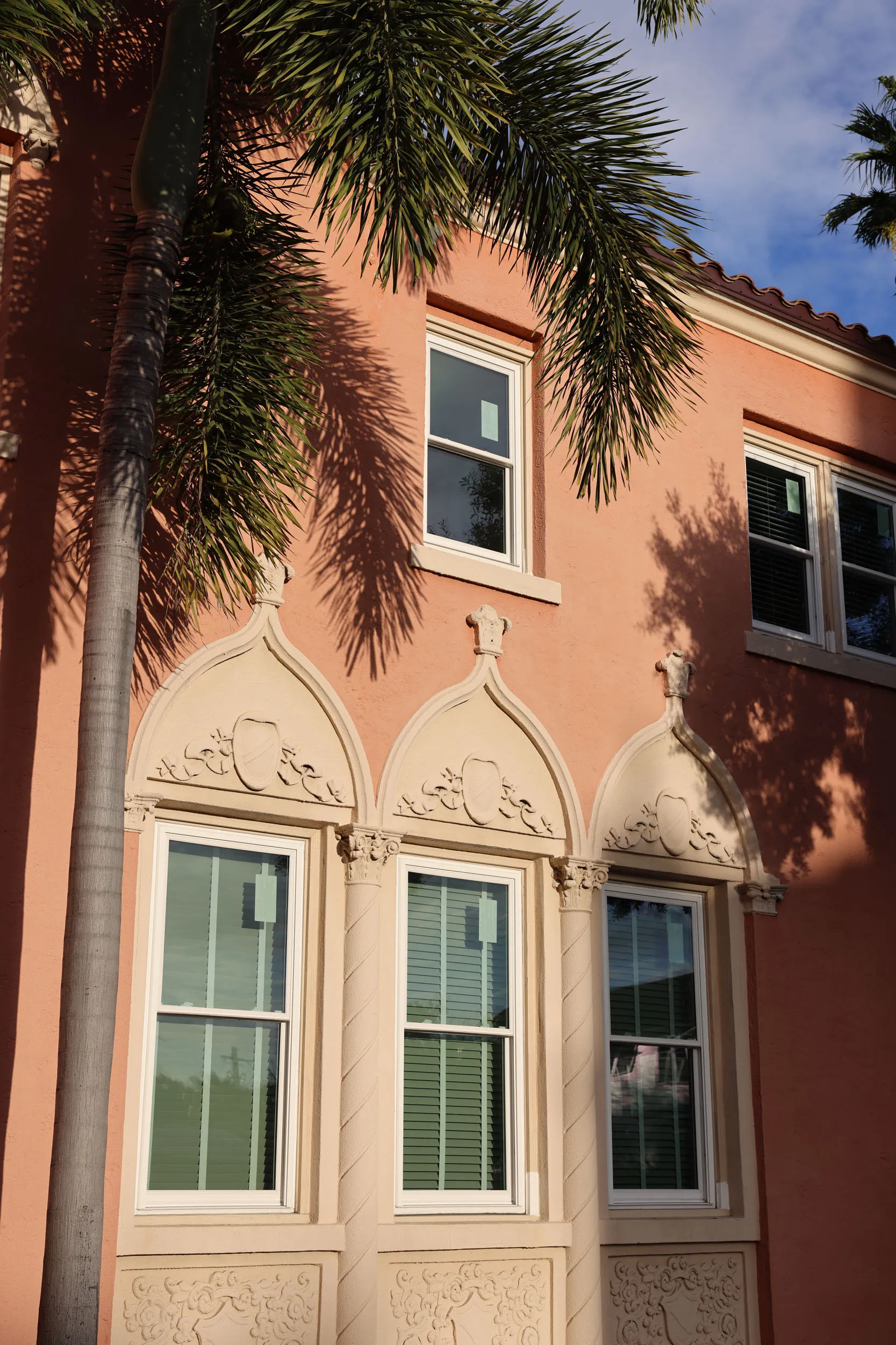 Close-up of a peach-colored building facade with three ornate arched windows on the lower level and two rectangular windows above, partially shaded by palm tree leaves.
